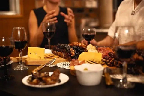 A group of friends sitting around a Charcuterie board that is filled up with Churchkhela and cheese and crackers. They all have glasses of red wine.