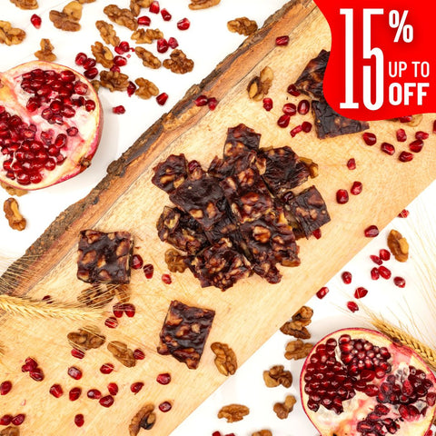 Squares of Pomegranate Walnut Chella FruityNuts Bites on a wooden board with pomegranates, wheat stalks, and a “15% off” label in the corner. Rustic and appetizing.