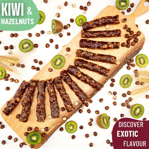 Slices of glossy Kiwi and hazelnut Chella Bar arranged in a fan shape on a wooden board, surrounded by kiwi halves, hazelnuts, and wheat stalks.