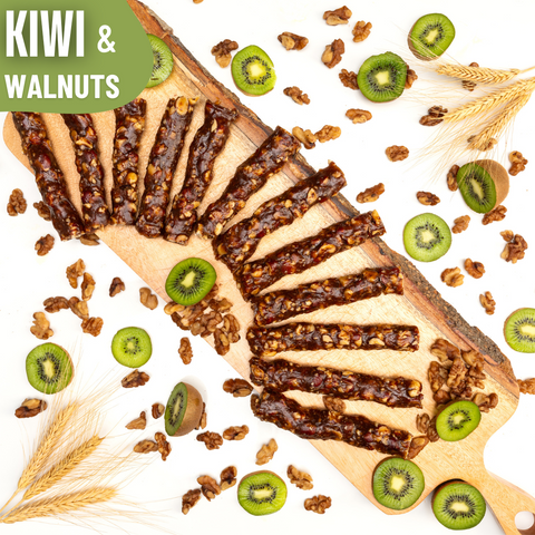Slices of glossy kiwi and walnut Chella Bar arranged in a fan shape on a wooden board, surrounded by kiwi halves, walnut pieces, and wheat stalks.