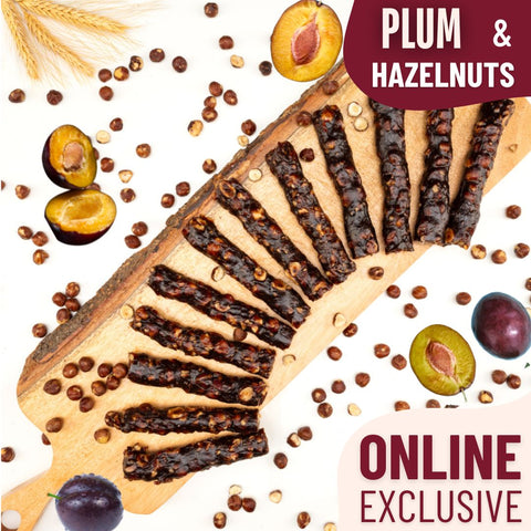 Slices of glossy plum and hazelnut Chella Bar arranged in a fan shape on a wooden board, surrounded by whole plums, hazelnuts, and wheat stalks.