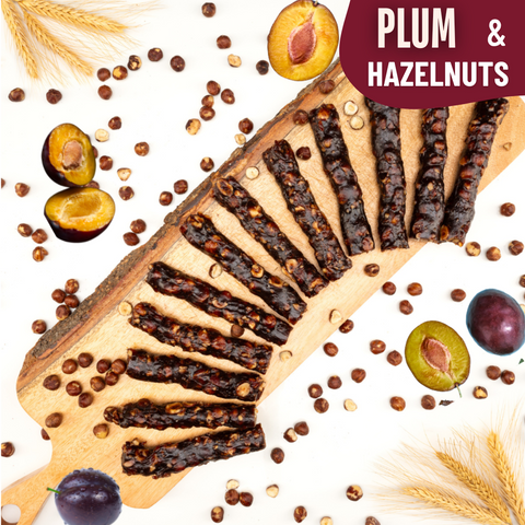 Slices of glossy plum and hazelnut Chella Bar arranged in a fan shape on a wooden board, surrounded by whole plums, hazelnuts, and wheat stalks.