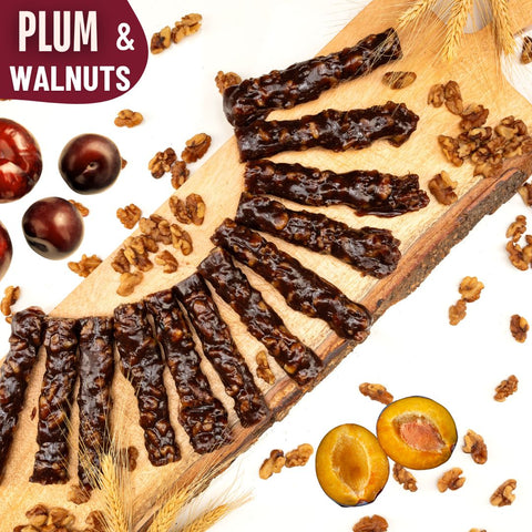 Slices of glossy plum and walnut Chella Bar arranged in a fan shape on a wooden board, surrounded by whole plums, walnut pieces, and wheat stalks.