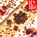 Squares of Pomegranate Walnut Chella FruityNuts Bites on a wooden board with pomegranates, wheat stalks, and a “15% off” label in the corner. Rustic and appetizing.